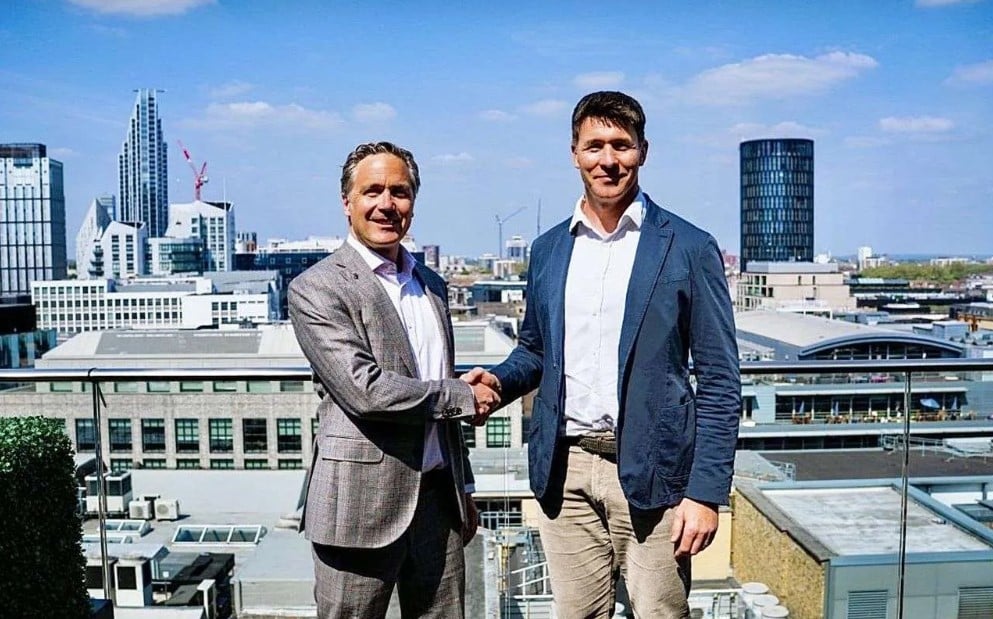 two white men in suits shake hands on a rooftop with a cityscape behind them