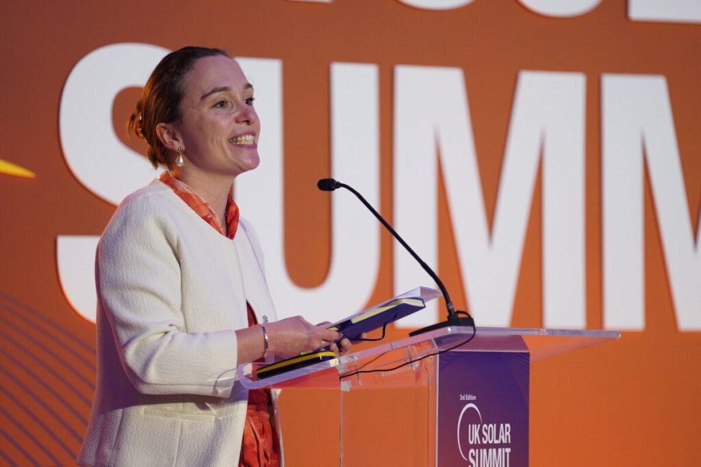 a woman speaks at a podium with a uk solar summit banner behind her