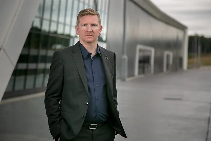 a white man in a black suit stands in front of a glass building