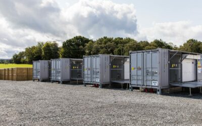 Green trees against three grey battery energy storage systems on gravel.