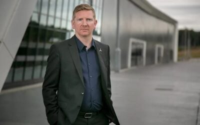 a white man in a black suit stands in front of a glass building