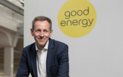 a white man in a suit gives a small smile as he sits in front of a wall with the good energy logo
