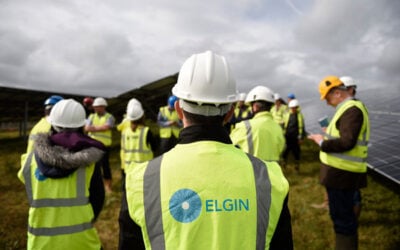 A man in an Elgin-branded high vis vest stands with his back to the camera, surrounded by other construction workers