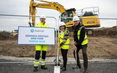 Three people stand in front of a large digger holding a sign announcing the greener grid project, spades, and pitchforks.