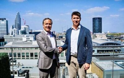 two white men in suits shake hands on a rooftop with a cityscape behind them